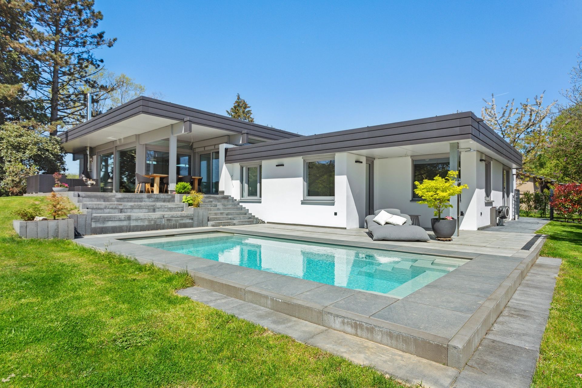 Moderner Bungalow mit Pool, großzügiger Steinterrasse und gepflegtem Rasen, umgeben von Bäumen.