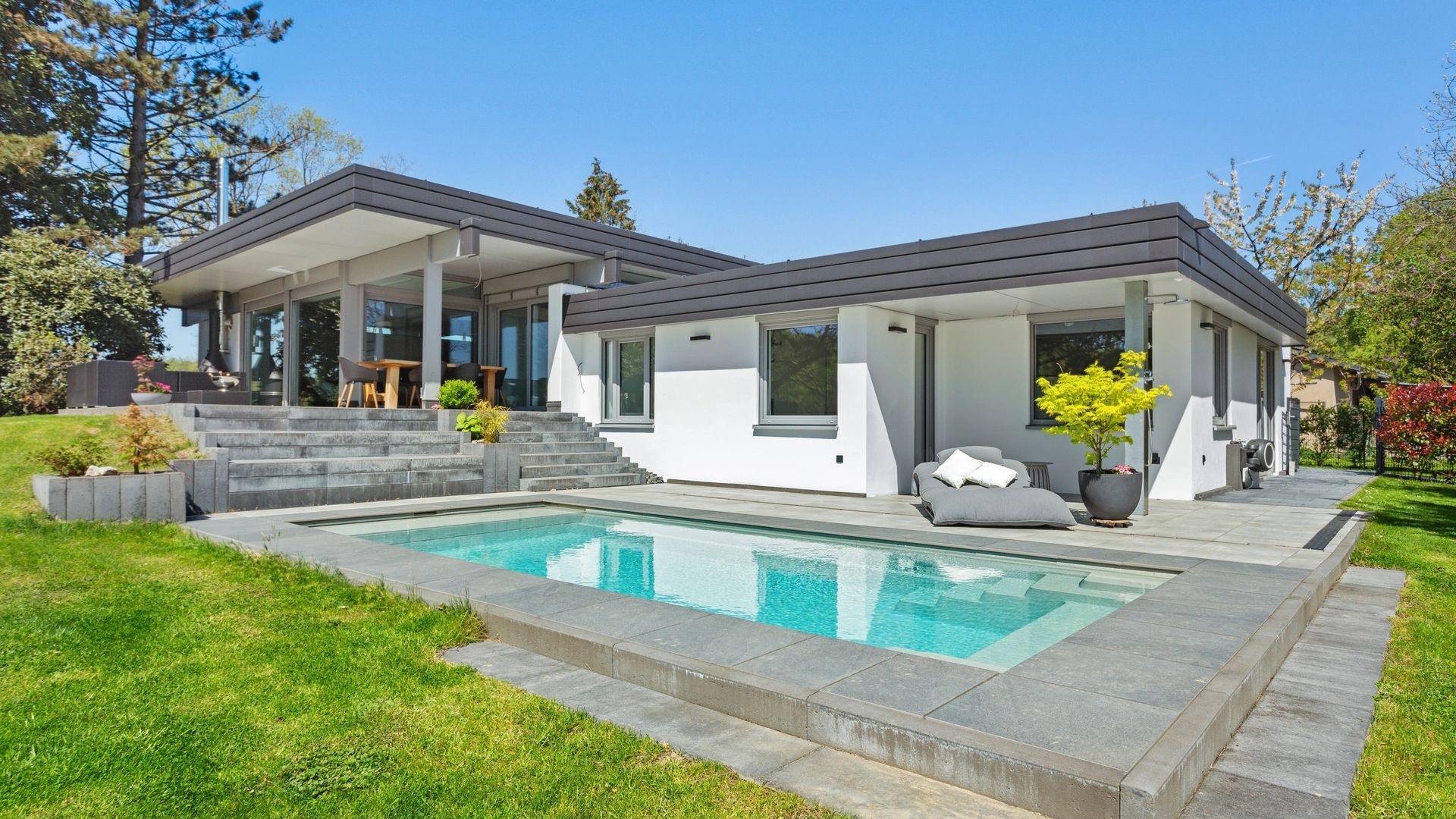 Moderner Bungalow mit Pool, großzügiger Steinterrasse und gepflegtem Rasen, umgeben von Bäumen.